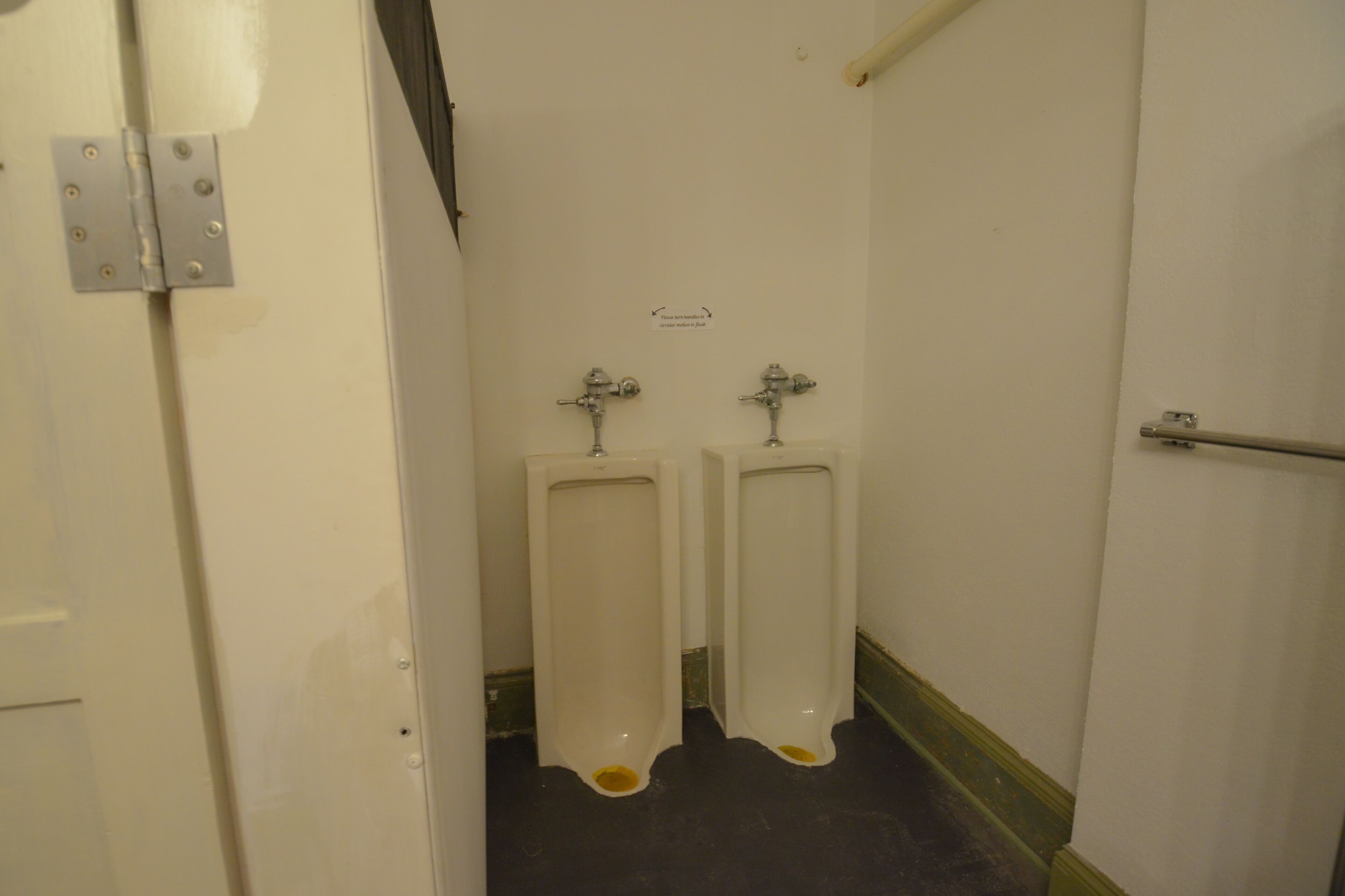 A men's restroom with two urinals, a white door, and a sign indicating the direction to the ladies' room.