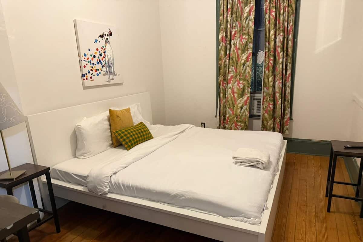 A neatly made bed with white linens, a green and yellow checkered pillow, and a white towel folded at the foot of the bed. There is a small table with a lamp on the left side of the room.