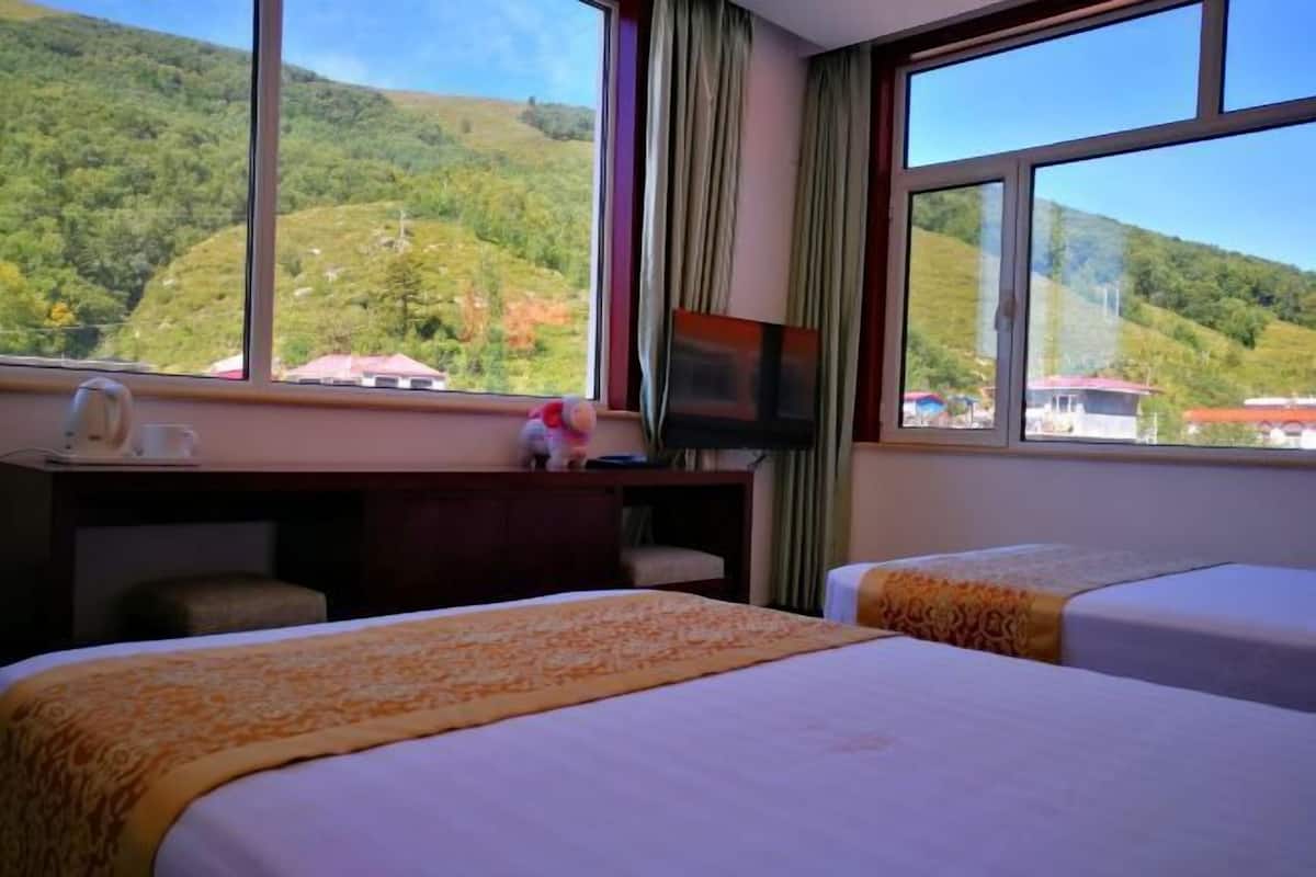 Habitación de hotel con dos camas, un escritorio y televisión, y una ventana con vistas a colinas verdes.