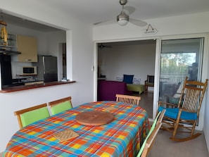 Une pièce comprenant un lit, un fauteuil à bascule en bois, une table à manger, un ventilateur de plafond et un coin cuisine visible à travers une porte ouverte.