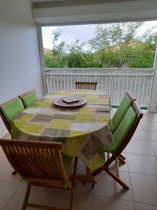 A round table with a checkered tablecloth, surrounded by wooden chairs with green cushions, on a balcony with a white railing and a view of greenery.