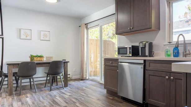 A modern kitchen with dark wooden cabinets, a built-in microwave, and a dishwasher. There is a dining area with a wooden table and chairs, and a window with a view of the outdoors.