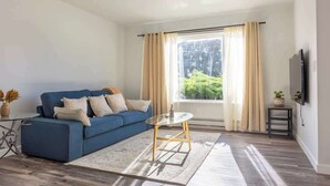 A living room with a blue sofa, a glass coffee table, a small side table, and a TV mounted on the wall.
