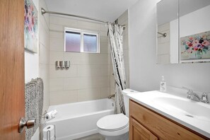 Une salle de bain avec une baignoire, des toilettes et un lavabo. Il y a une fenêtre au-dessus de la baignoire, un rideau de douche et un tableau au mur.