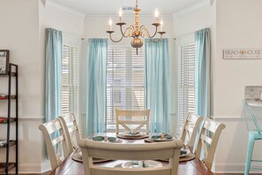 Une salle à manger avec une table en bois dressée pour six personnes, entourée de chaises, d’un lustre et de rideaux bleu clair.
