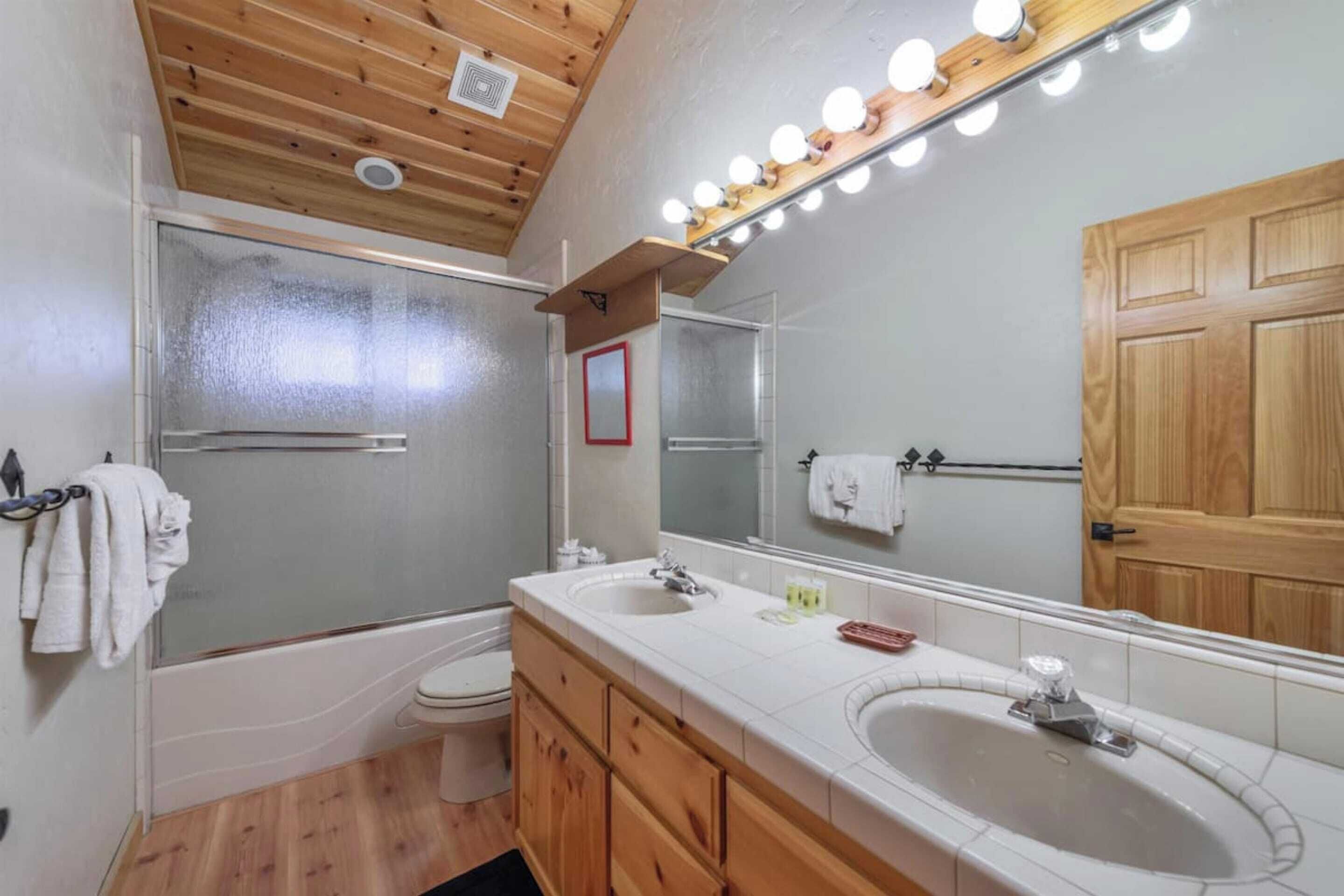 A bathroom with a wooden ceiling, a large mirror with lights, a bathtub, and a sink.