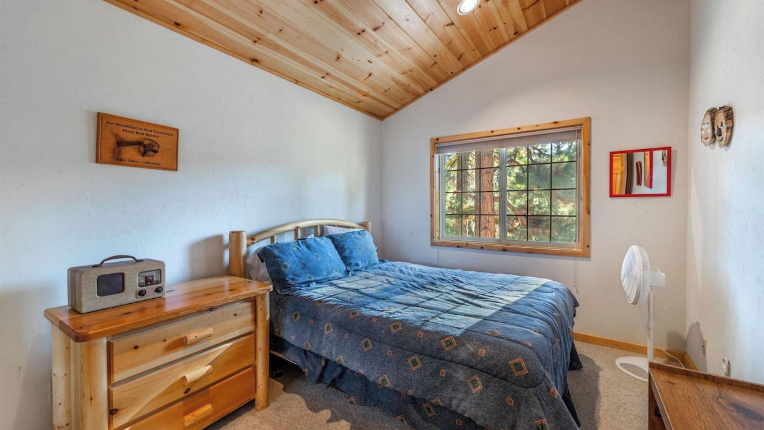 A bedroom with a wooden ceiling, a bed with blue bedding, a wooden dresser, a window with a view of trees, and a wall-mounted radio.