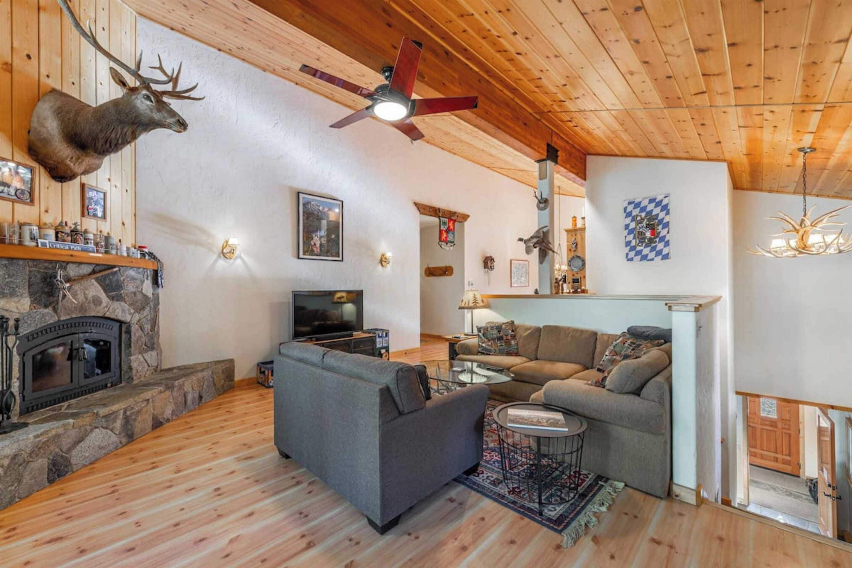 Una sala de estar acogedora con chimenea de piedra, cabeza de venado montada, ventilador de techo y televisor.