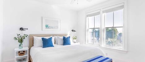 Een lichte, witte slaapkamer met een groot bed, een bankje, een plafondventilator en een raam met jaloezieën.