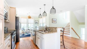 A spacious kitchen with white cabinetry, a granite countertop, and a central island. There are hanging pendant lights and a staircase leading to an upper level. - Sea Shell by Avantstay Breezy Beachside Getaway w/ Rooftop Deck & 6 BRs (Isle of Palms)