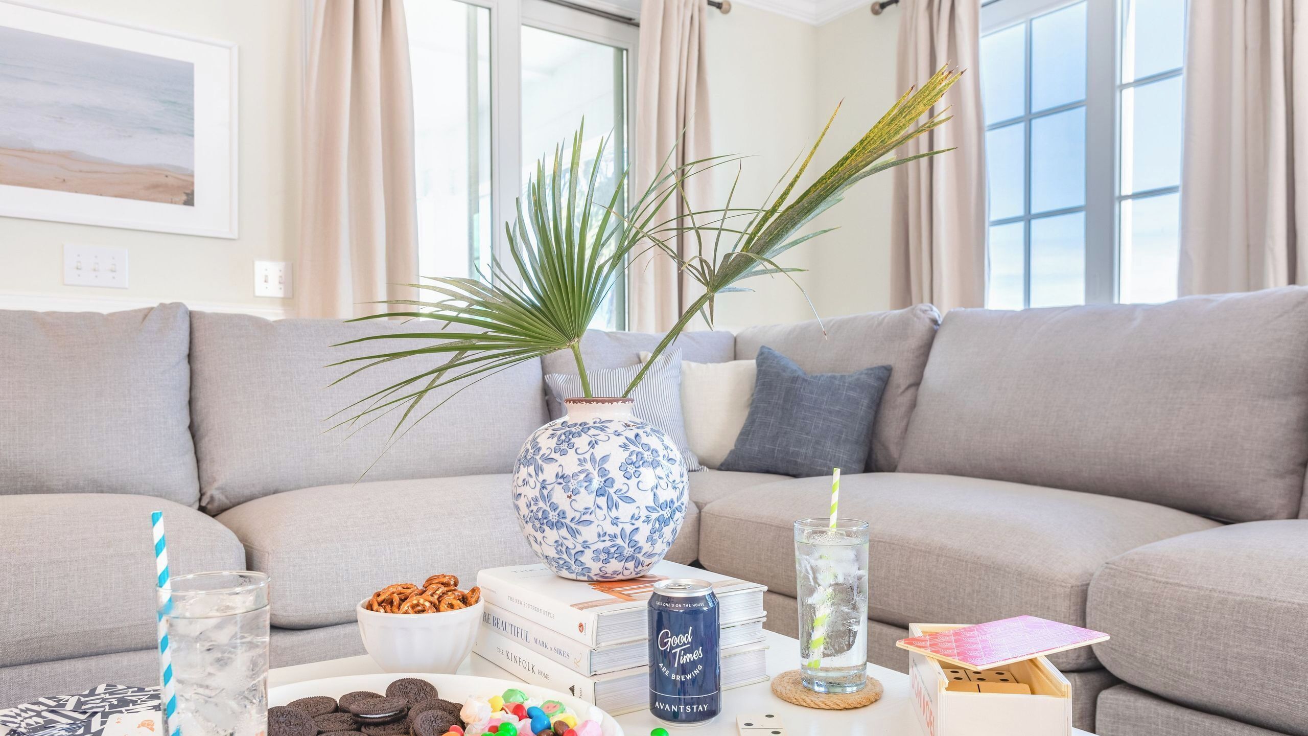 A living room with a sofa, a coffee table with snacks, books, a vase with plants, and a glass of water.