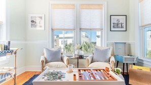 A room with two armchairs, a coffee table with a puzzle, a tray with a teapot, and a view of buildings through large windows. - Sea Shell by Avantstay Breezy Beachside Getaway w/ Rooftop Deck & 6 BRs (Isle of Palms)