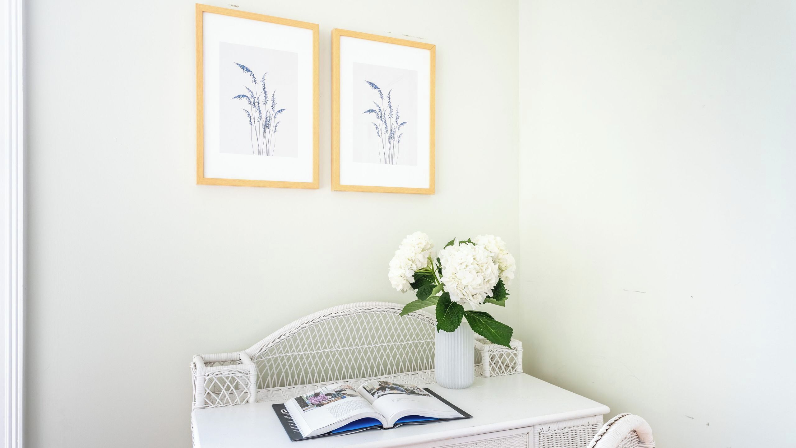 A white wicker chair, a white table with a book and a vase of white flowers, and two framed botanical illustrations on the wall.