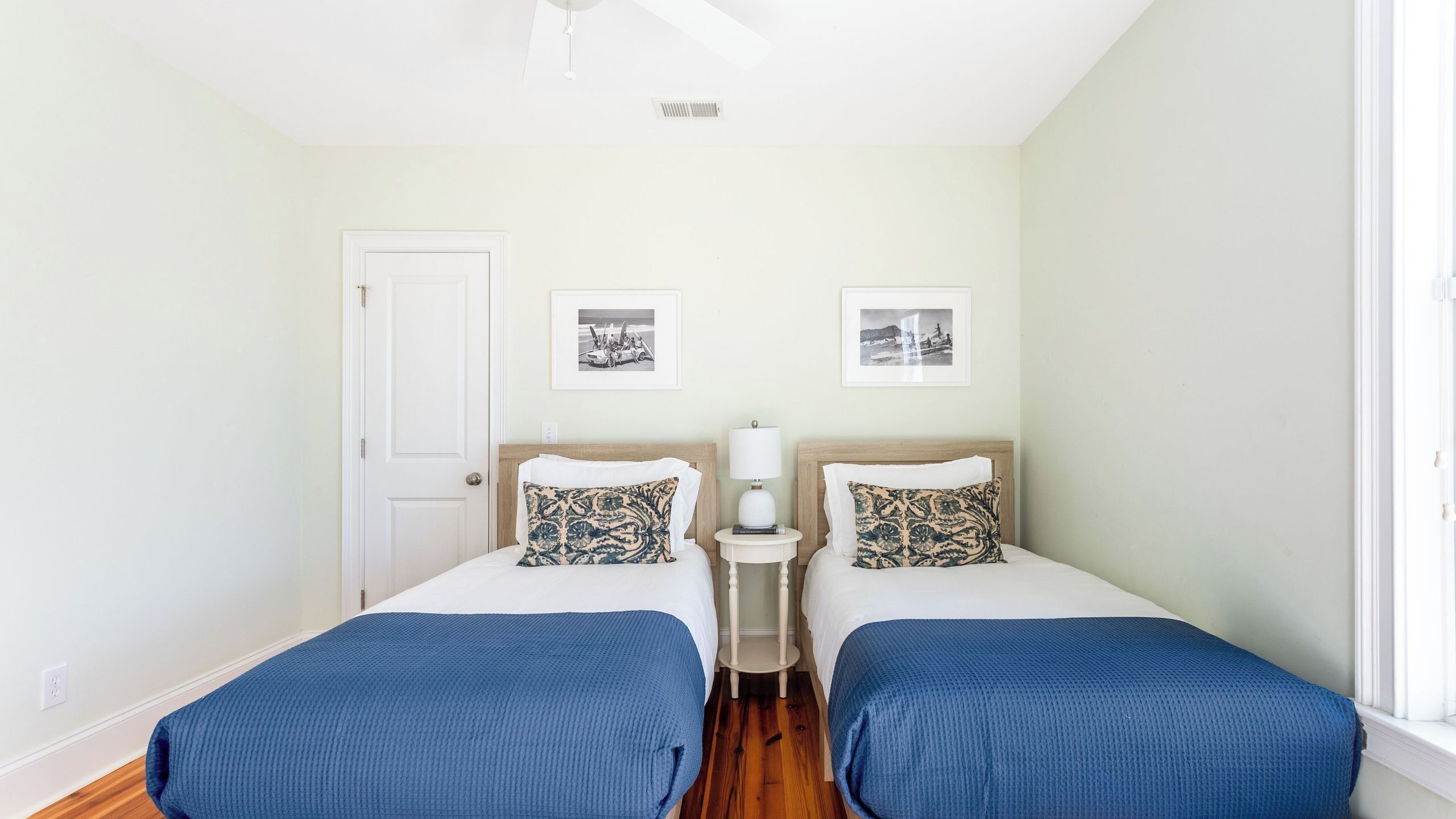 Une chambre avec deux lits, une porte blanche, une petite table de chevet avec une lampe, et deux tableaux encadrés au mur.