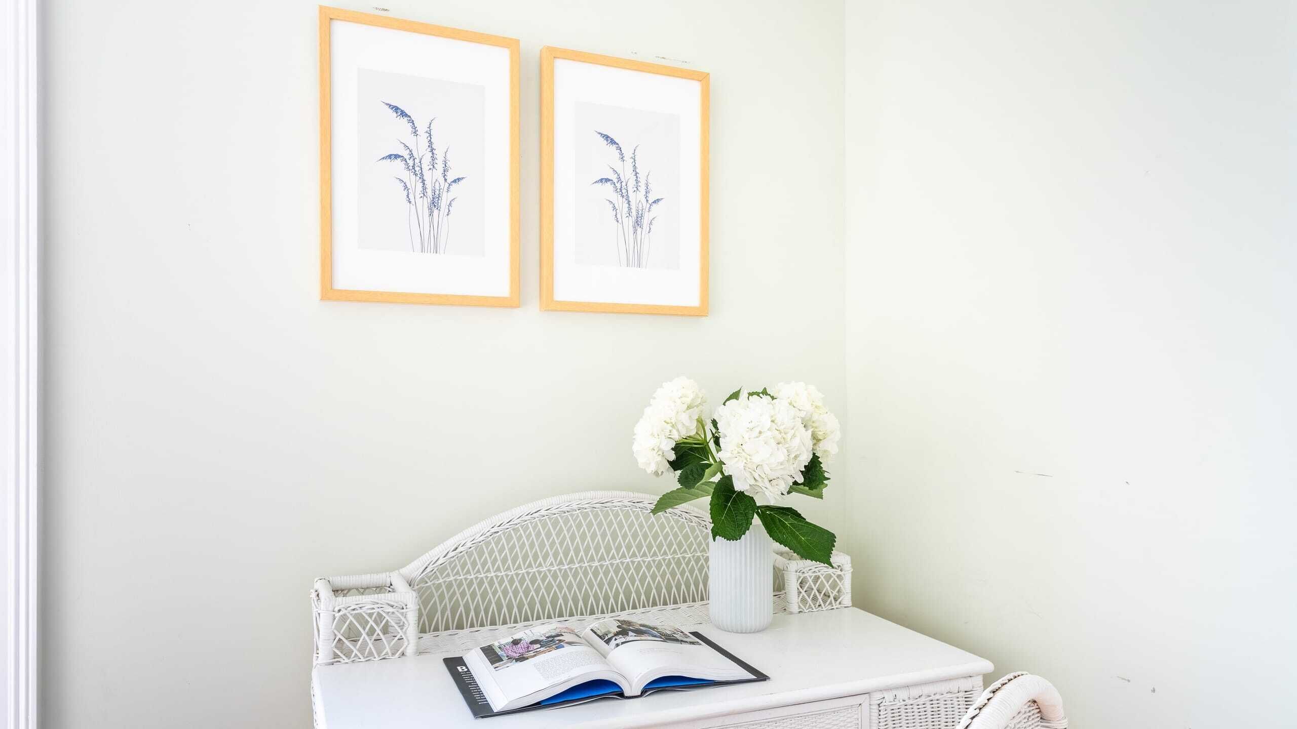 A room with a white wicker chair, a white table with a vase of white flowers, and two framed botanical illustrations on the wall.