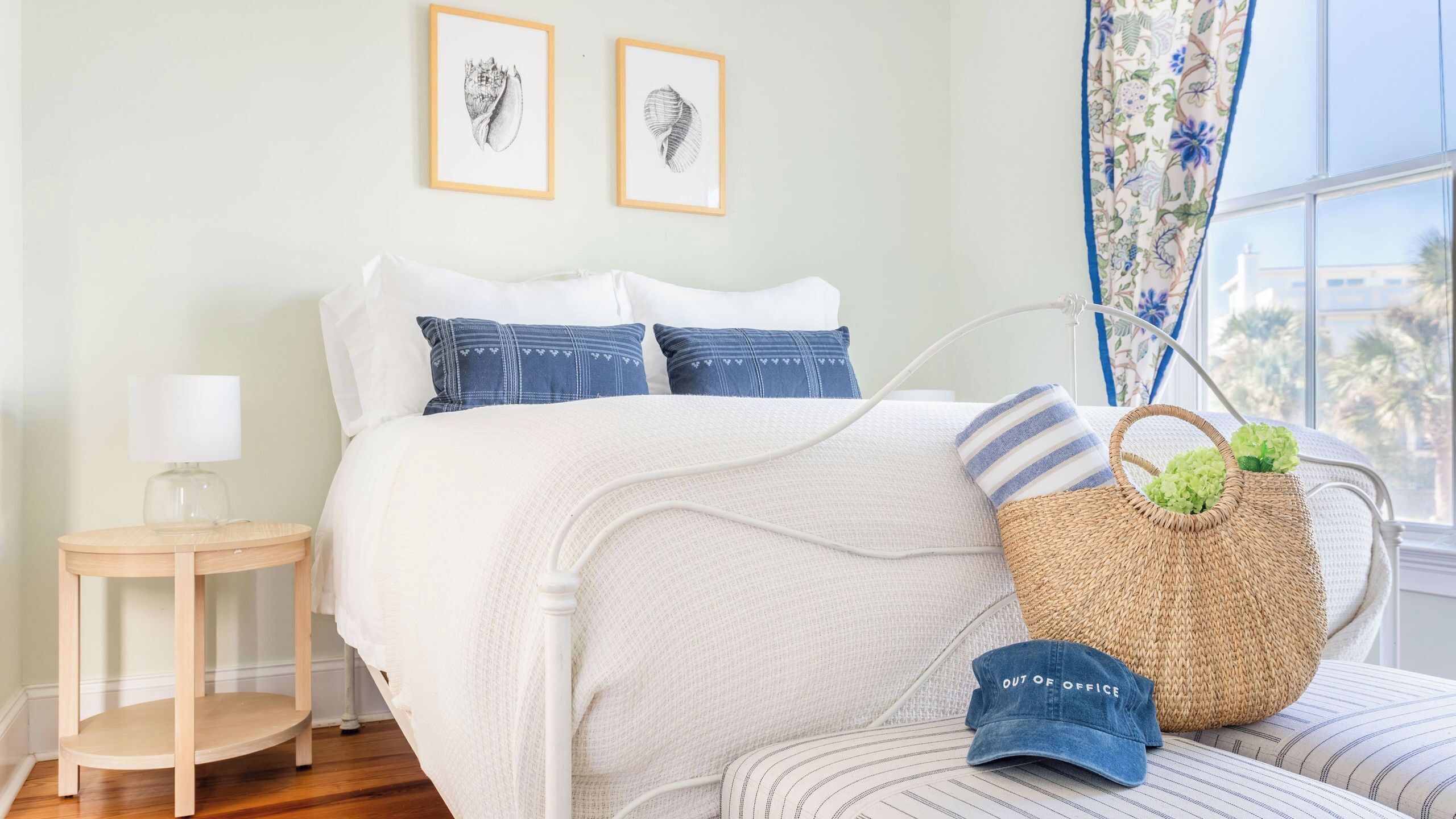 A bedroom with a bed, pillows, a bedside table, a lamp, a window with curtains, and two framed pictures on the wall.