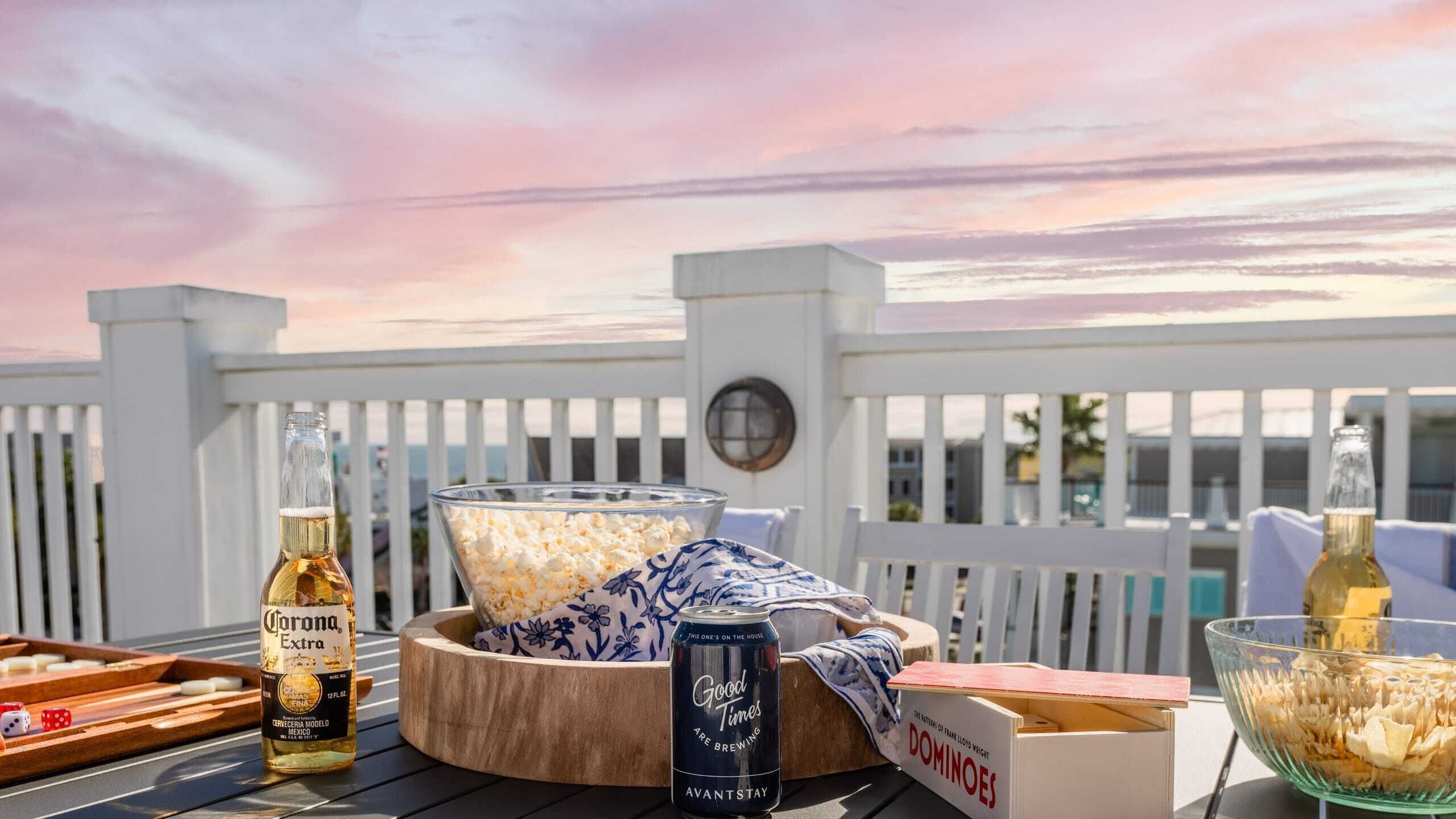 Een tafel gedekt met een fles Corona Extra bier, een blikje Good Times bier, een schaal popcorn en een doos Domino's pizza op een terras met een witte omheining en een zonsondergang op de achtergrond.