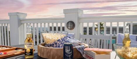Une table en bois sur laquelle se trouvent une bouteille de bière Corona, une canette de bière Good Times, un bol de pop-corn et une boîte de jeux de dominos, le tout placé sur une terrasse avec une clôture blanche et un coucher de soleil en arrière-plan.
