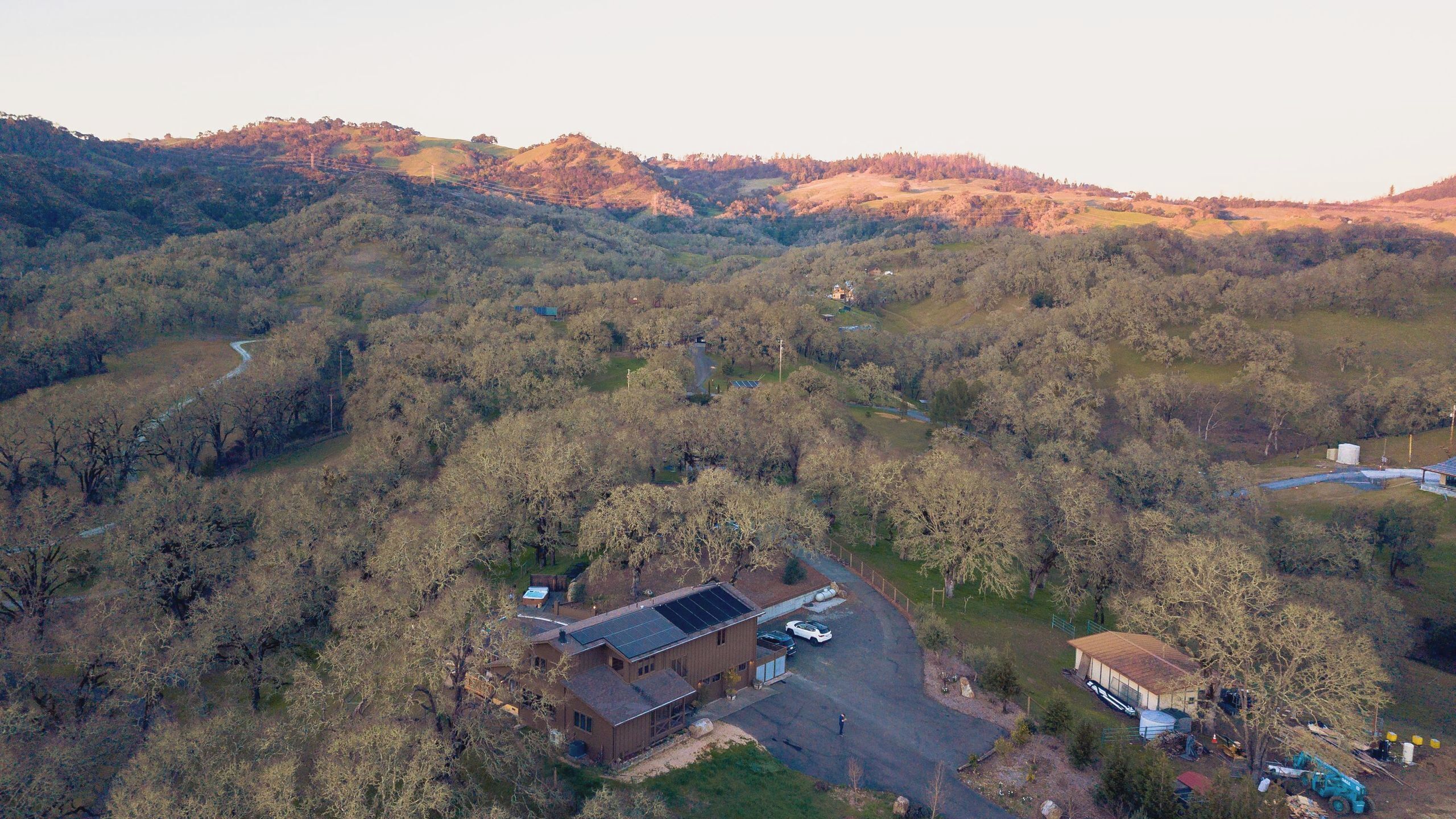 Vista aérea de uma propriedade com um edifício grande, painéis solares e uma área de estacionamento, tudo cercado por uma floresta densa.