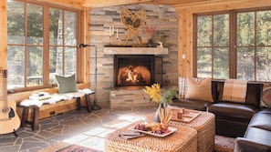 A cozy cabin interior with a stone fireplace, wooden furniture, and a view of the forest.