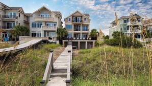A beachfront property with multiple-story houses, a wooden boardwalk leading to the beach, and a clear sky. - Oceanfront, Pool+hot Tub, Game Room, Views Steps to Beach Lido by Avantstay (Hilton Head Island)