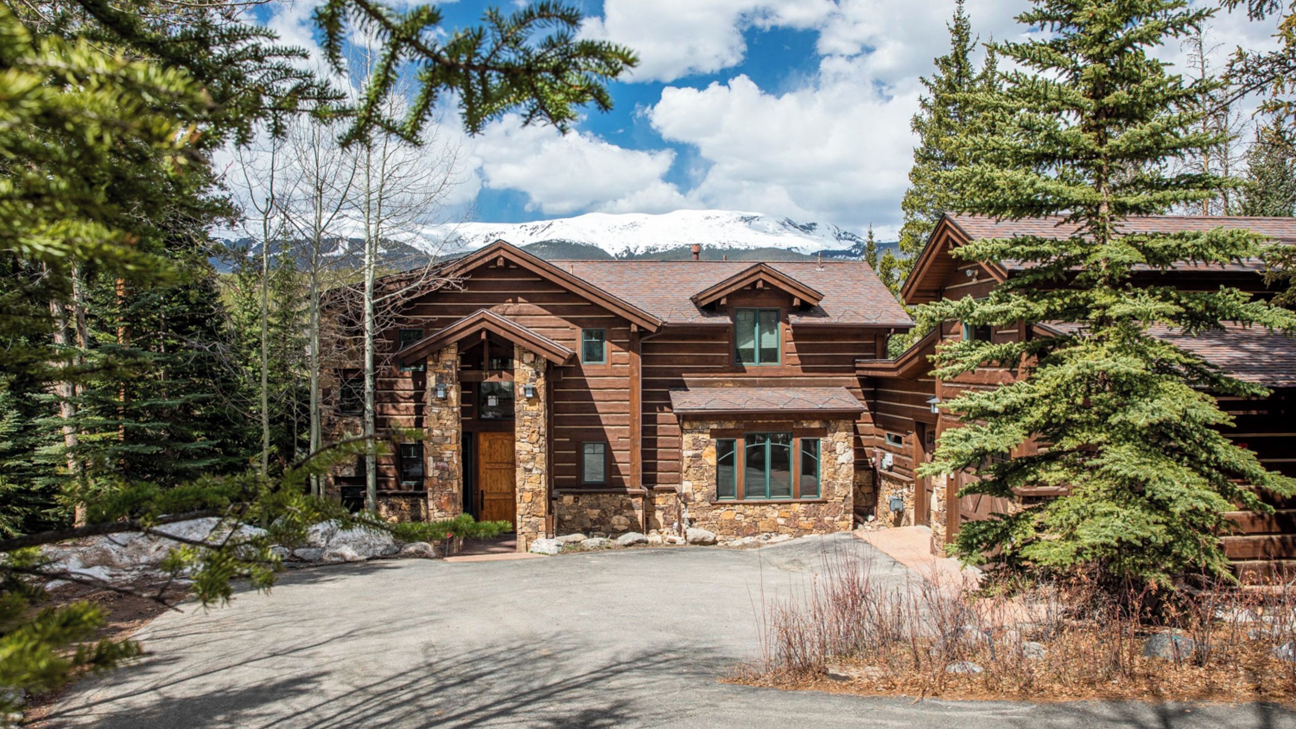 A log cabin with a stone foundation, a front porch, and a view of snow-capped mountains.
