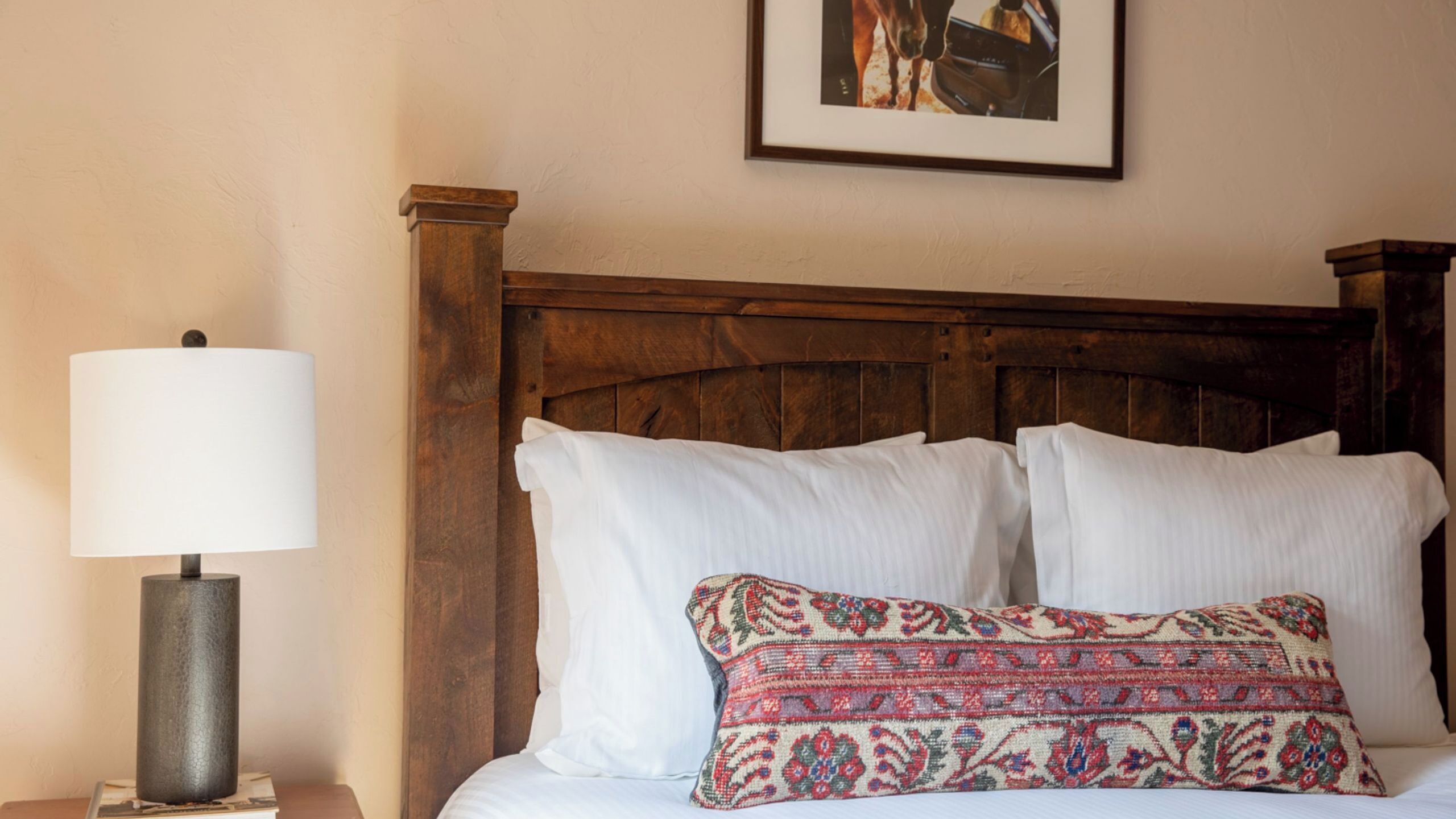 A bed with white linens, a patterned pillow, a wooden headboard, and a lamp on a bedside table.