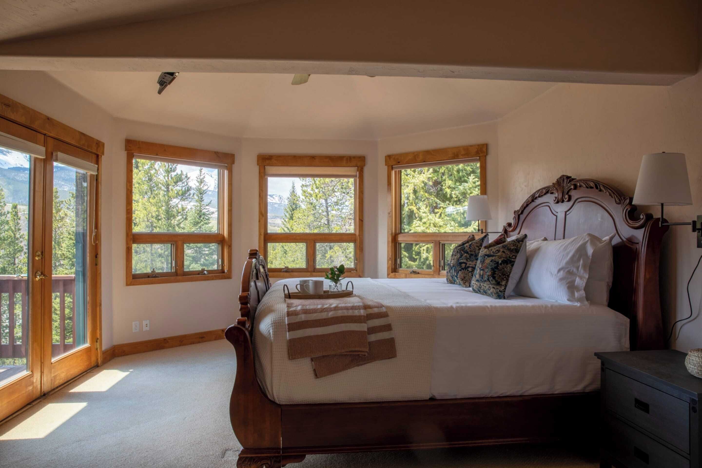 A bedroom with a large bed, wooden headboard, and multiple windows offering a view of trees and mountains.