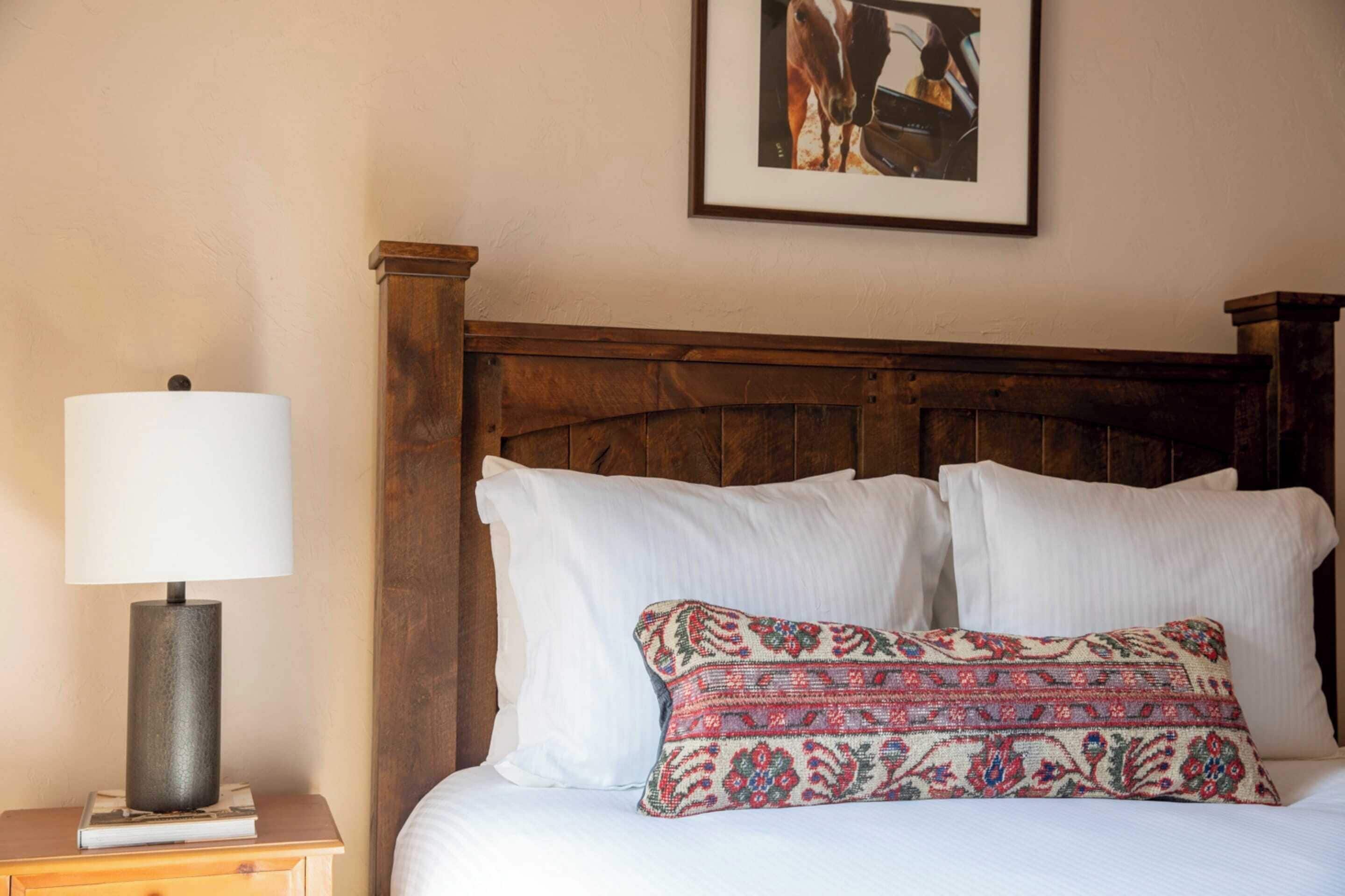 A neatly made bed with white linens and a patterned pillow, a wooden headboard, and a nightstand with a lamp and a book.