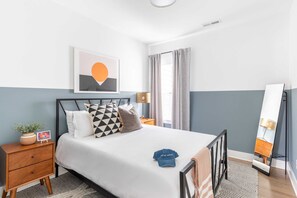 A neatly arranged bedroom with a bed, bedside tables, a nightstand, a mirror, and a wall-mounted artwork.