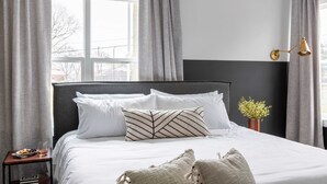 Una cama bien hecha con ropa blanca y un cojín de estampado geométrico, una mesita de noche con una copa de vino y aperitivos, y una pequeña planta en maceta en la mesita de noche.