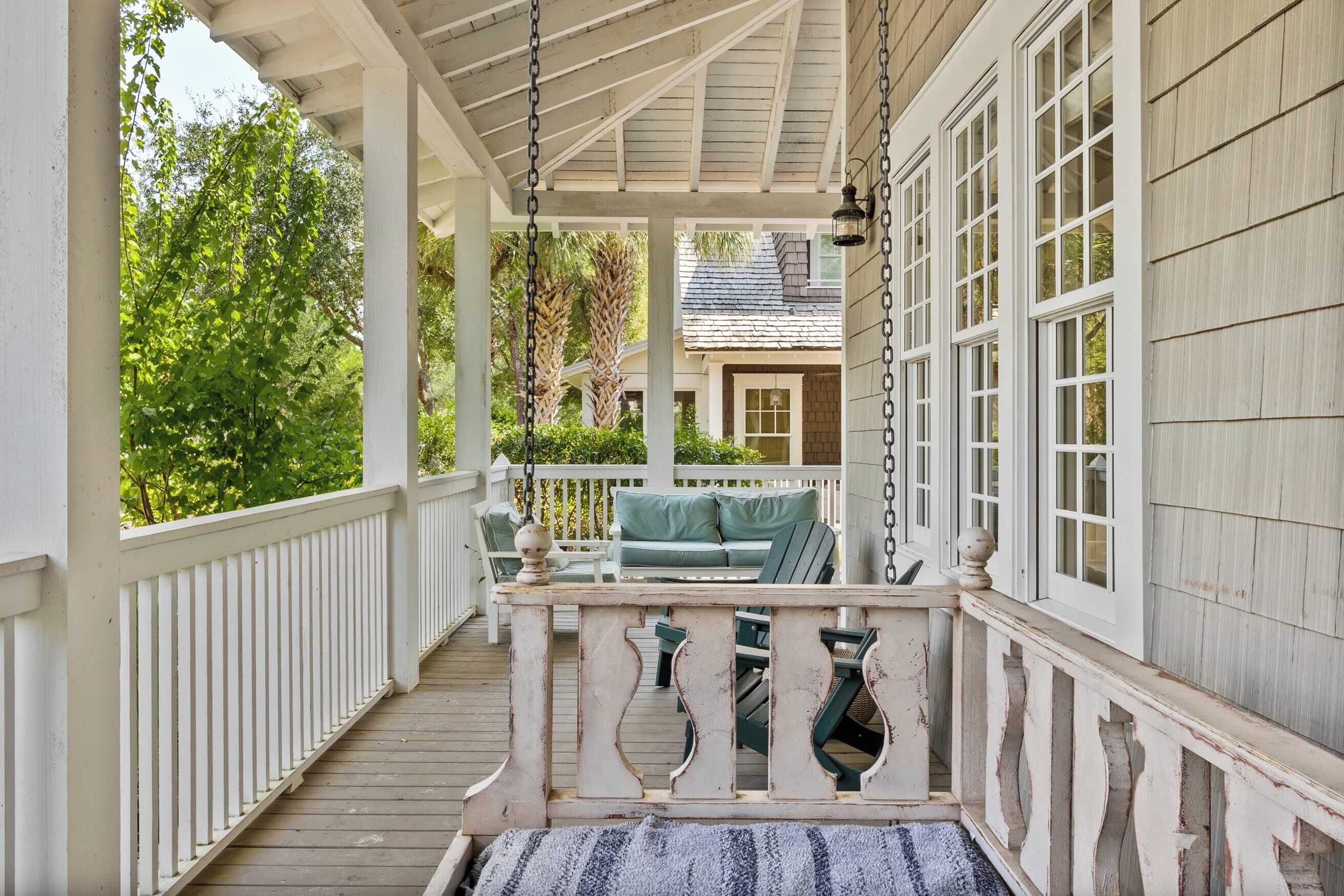 Une véranda couverte avec une balustrade blanche, des meubles d’extérieur verts et une vue sur une maison en arrière-plan.