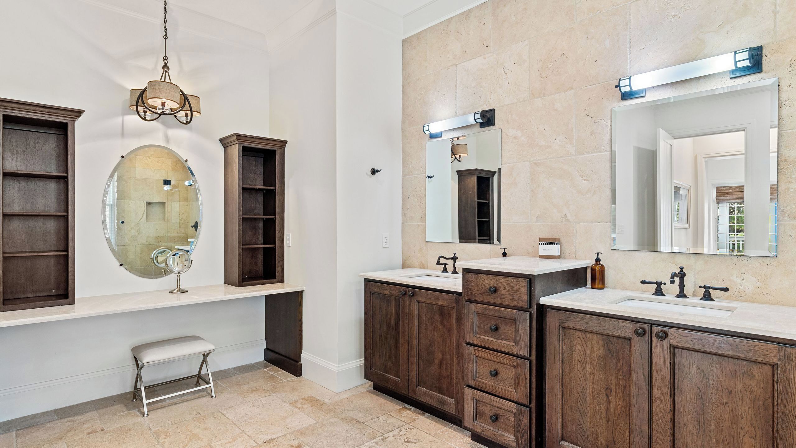 A bathroom with two sinks, wooden cabinets, a vanity, a round mirror, and a bench.