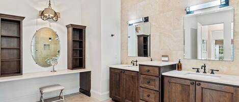 A bathroom with two sinks, wooden cabinets, a vanity, a round mirror, and a bench.