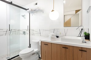 A modern bathroom with a glass shower enclosure, a white toilet, a wooden vanity with a sink, and a mirror above the vanity. - The Llama House by Avantstay Featured on Hgtv, Resort Style, Pool & Spa! Permit#4277 (Palm Springs)