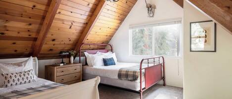 Un dormitorio acogedor en el ático, con techo inclinado, una cama, un aparador y una ventana con vistas a árboles.