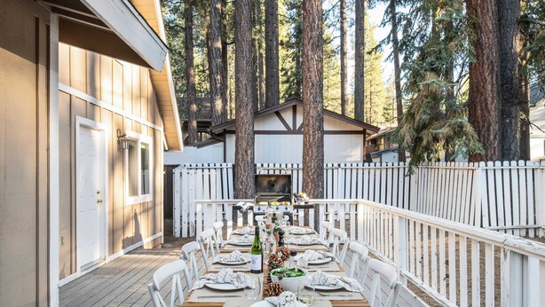An outdoor dining area with a wooden deck, white chairs, and a table set for a meal. - Hartwood by Avantstay Cozy Big Bear Abode w/ Spacious Deck & Stone Fireplace (Big Bear Lake)