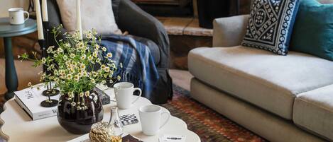 A cozy living room with a fireplace, a white sofa, a coffee table with books and a vase of flowers, and a patterned rug.