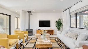 A modern living room with a fireplace, sofa, armchairs, and a large window. - Lava Rocks by Avantstay Gorgeous Desert Haven w/ Pool, Hot Tub, Fire Pit & Ping Pong! (Yucca Valley)