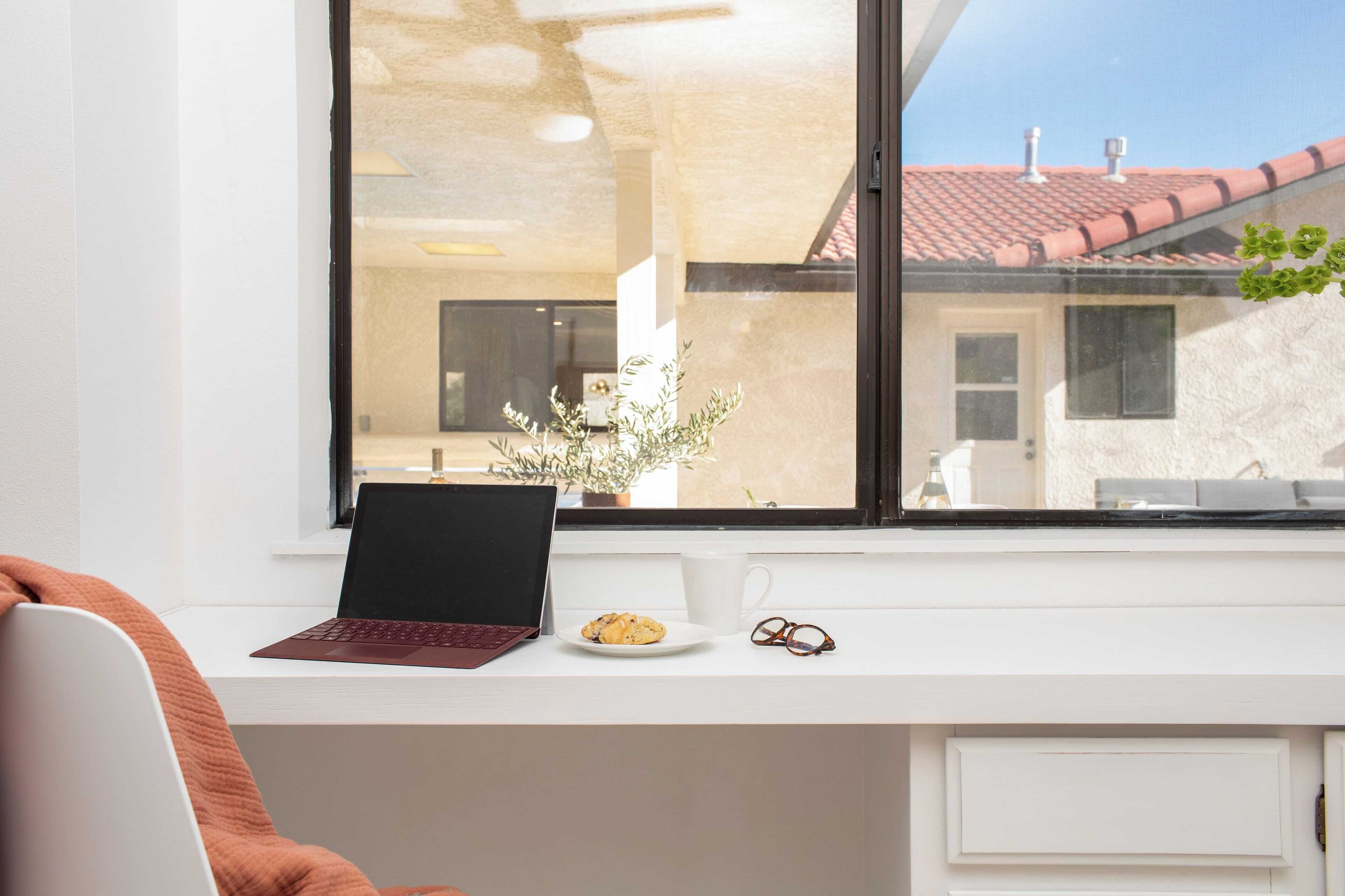 A modern workspace with a laptop, a plate of food, and a pair of glasses on a white desk by a large window.