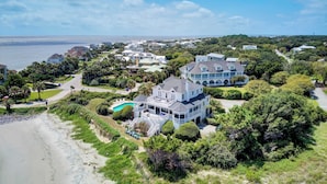 A large, multi-story house with a pool, surrounded by lush greenery and a beach.