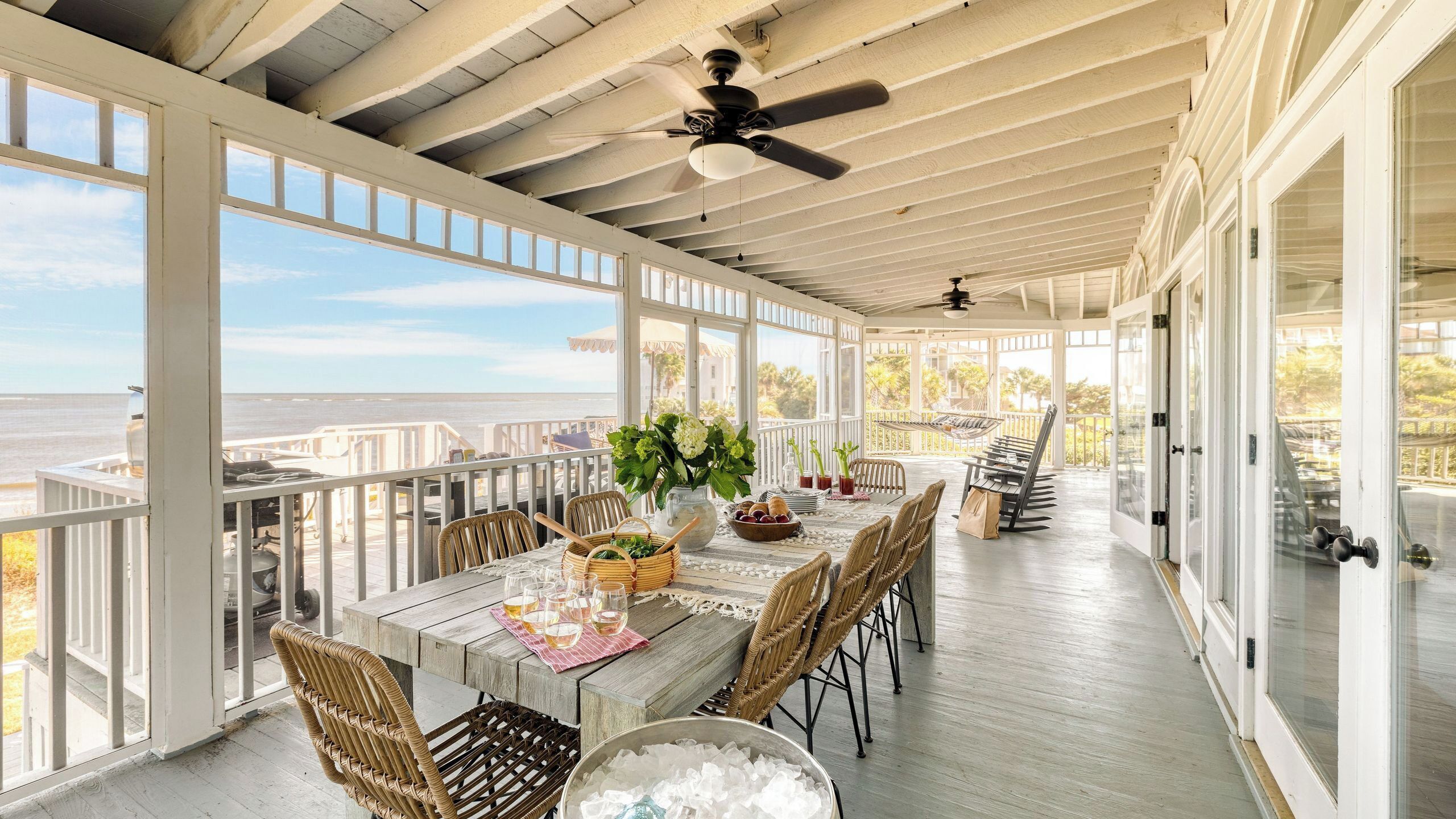 A covered outdoor dining area with a view of the sea, wooden tables, wicker chairs, and a ceiling fan.