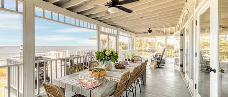 A covered outdoor dining area with a view of the sea, wooden tables, wicker chairs, and a ceiling fan.