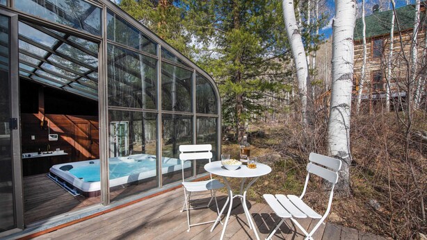 A glass-enclosed hot tub area with a wooden deck, white chairs, and a view of a cabin in a forest. - Condor by Avantstay Gorgeous Mountain Home w/ Hot Tub & Sauna (Vail)