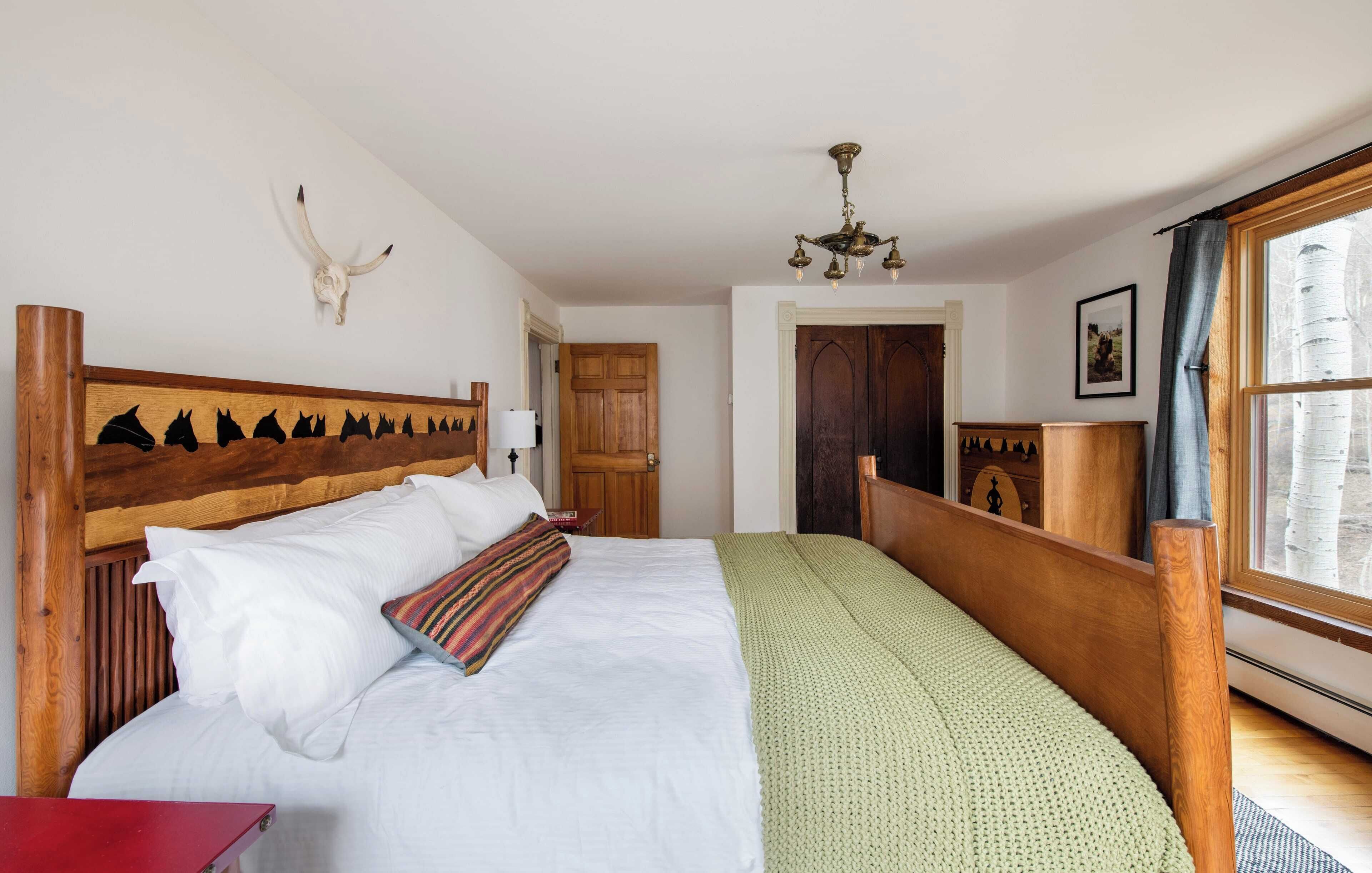 A bedroom with a wooden headboard, a large bed with white and green bedding, a mounted cow skull, a chandelier, and a window with a view of birch trees.
