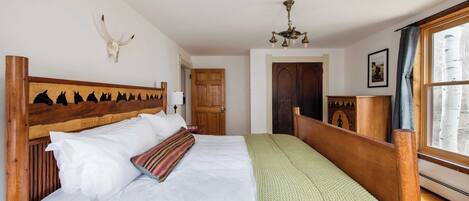 A bedroom with a wooden headboard, a large bed with white and green bedding, a mounted cow skull, a chandelier, and a window with a view of birch trees.