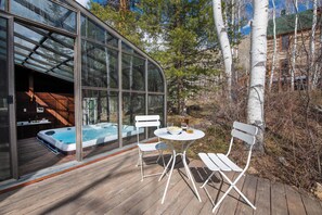 A glass-enclosed hot tub area with a wooden deck, white chairs, and a wooden cabin in the background.