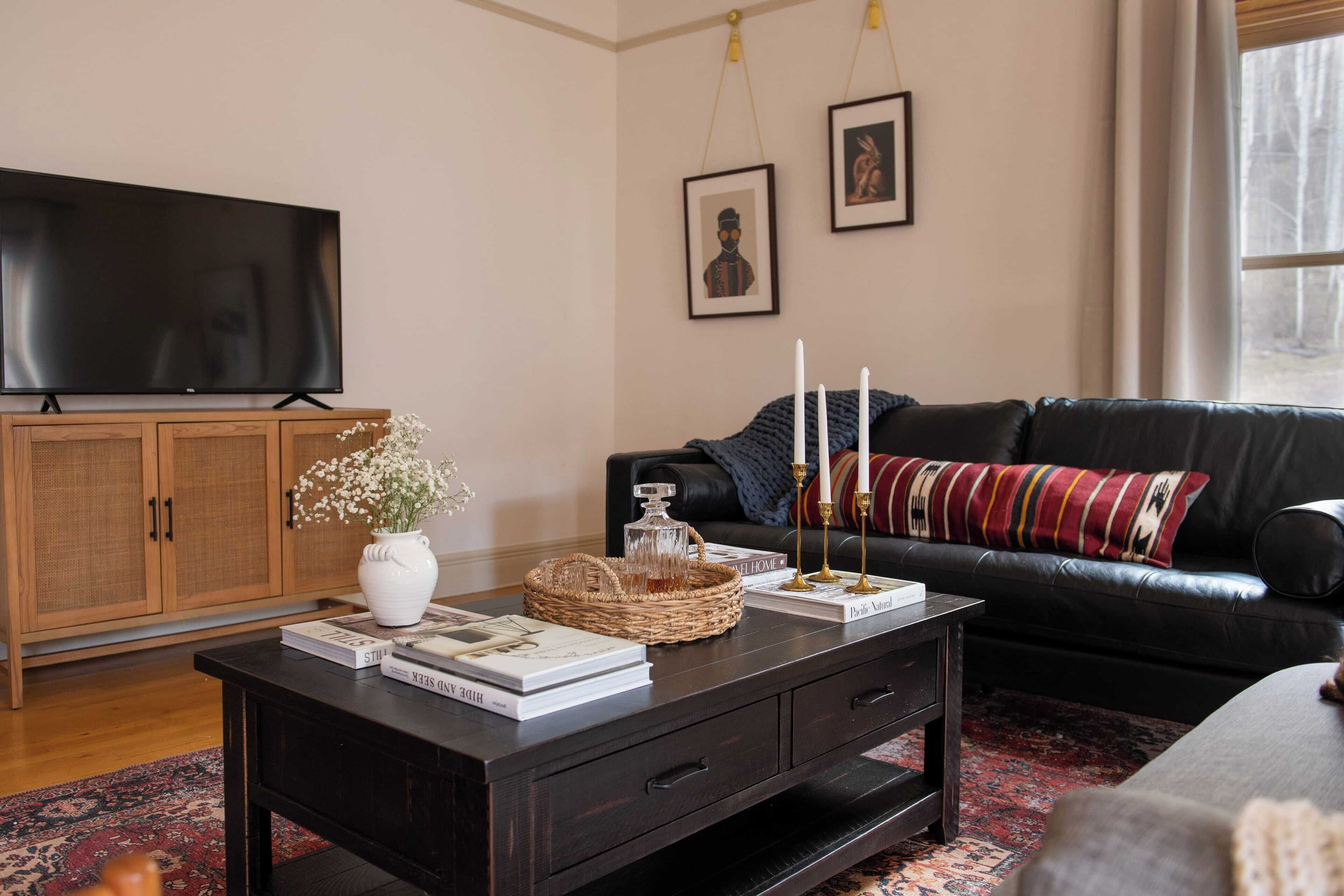 A living room with a dark wooden coffee table, a black leather sofa, a television, and a large window with curtains.