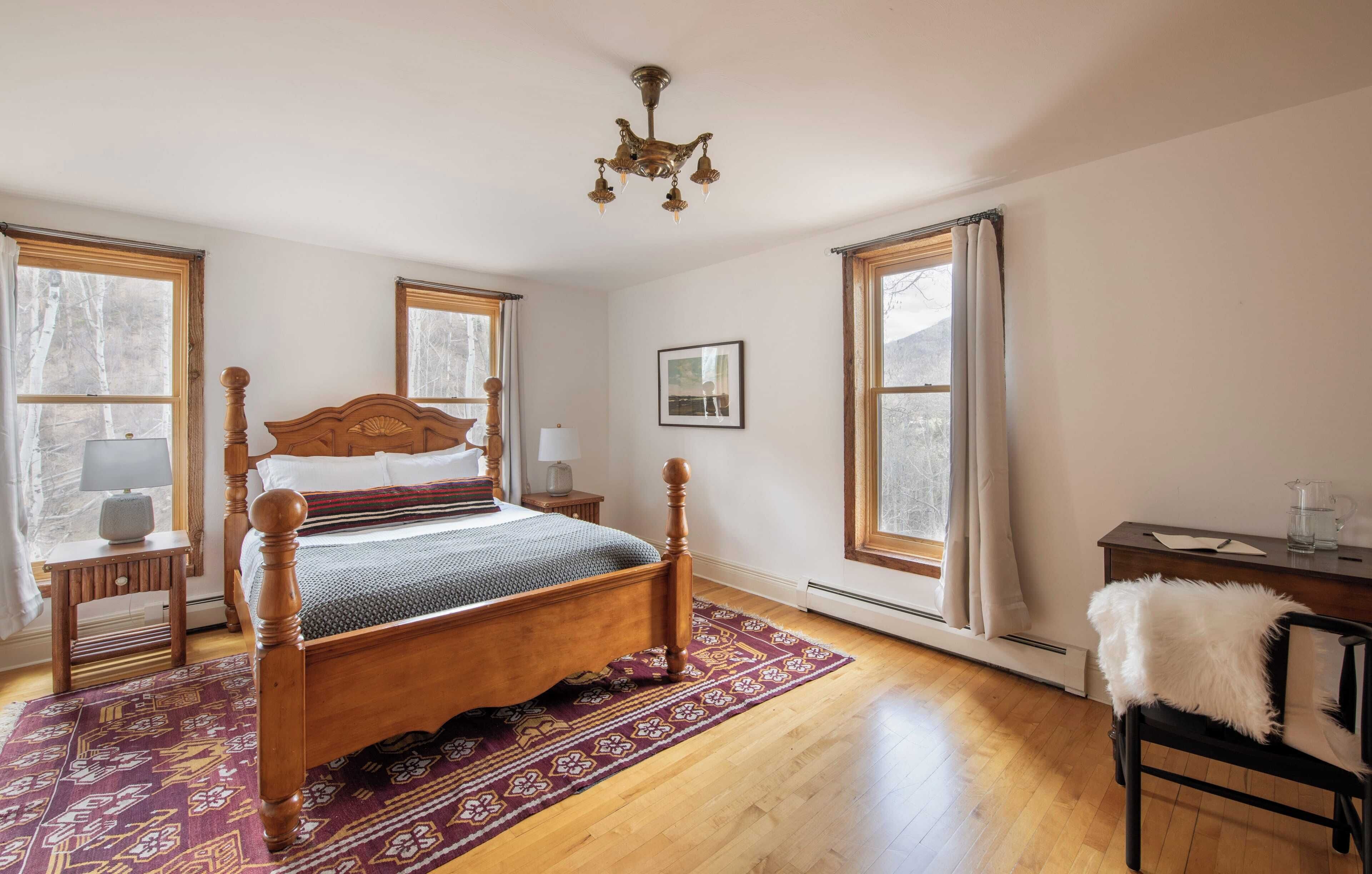 Une chambre à coucher avec un lit en bois, un tapis à motifs, un bureau avec une chaise et de grandes fenêtres.