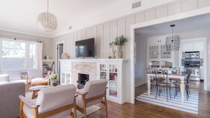 A living room with a fireplace, a television, and a dining area with a kitchen in the background. - Topaz By Avantstay Adorable Cottage Walking Distance to Beach w/ Large Patio (La Jolla)